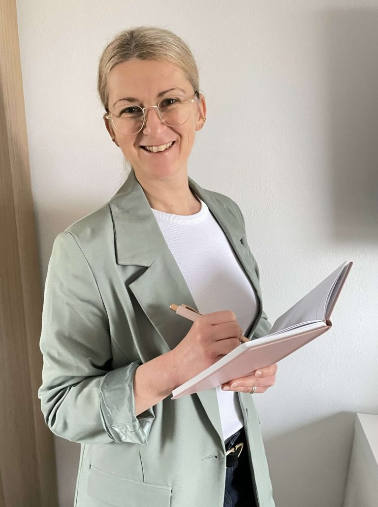 Isabella Örtülü Eine lächelnde Frau mit Brille hält ein Notizbuch und einen Stift in der Hand.