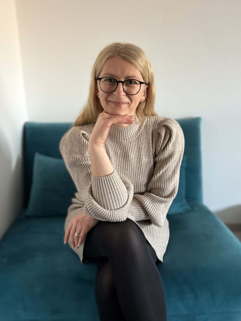 Isabella Örtülü Frau mit langen blonden Haaren sitzt auf einem blauen Sofa und lächelt freundlich.